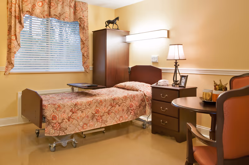 A furnished hospital-style bedroom with a floral bedspread, dresser, lamp, and window curtains.