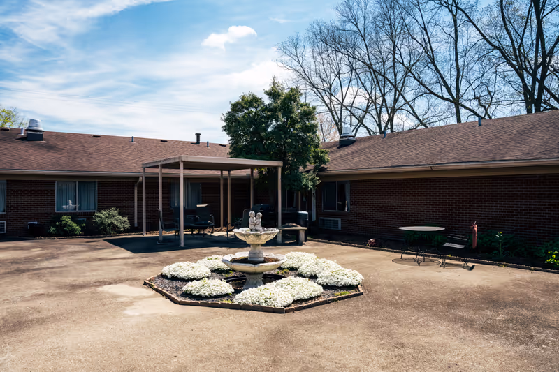 Brick senior living building with a paved courtyard, central fountain, and outdoor seating under a canopy.