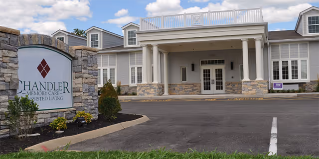 Exterior view of a senior living facility with a stone and white column entrance. A sign in the foreground reads 'Chandler Memory Care Assisted Living'. The building has multiple windows and dormers on the roof, with a clear sky and some clouds above.