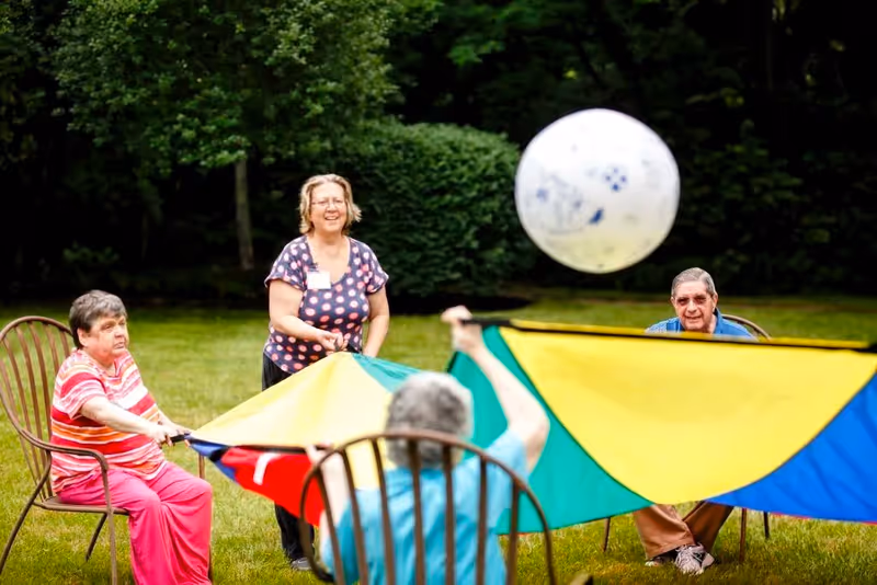 Four elderly individuals sitting in chairs outside on grass, playing with a colorful parachute and a large white ball, surrounded by green bushes and trees.