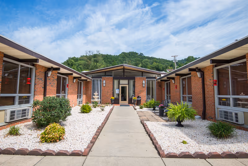Exterior view of Prestonsburg Health Care Center showing a pathway leading to the main entrance flanked by two single-story brick buildings with windows and air conditioning units. The landscaped area along the pathway features white gravel, green shrubs, and potted plants under a partly cloudy blue sky with a forested hill in the background.