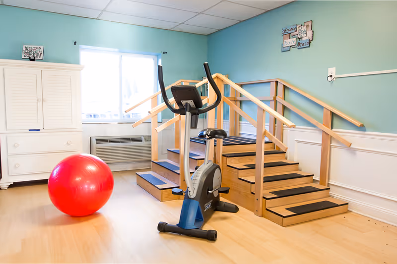 A rehabilitation room with a stationary exercise bike, a large red exercise ball, and a wooden staircase with handrails on both sides for physical therapy. The room has light blue walls, a window with natural light, and a white cabinet.