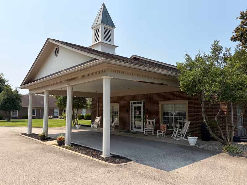 Brick facility entrance with a covered portico and white rocking chairs outside.