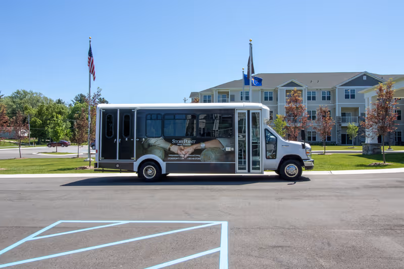 A StoryPoint shuttle bus parked in front of a multi-story senior living building with flags and landscaped grounds.