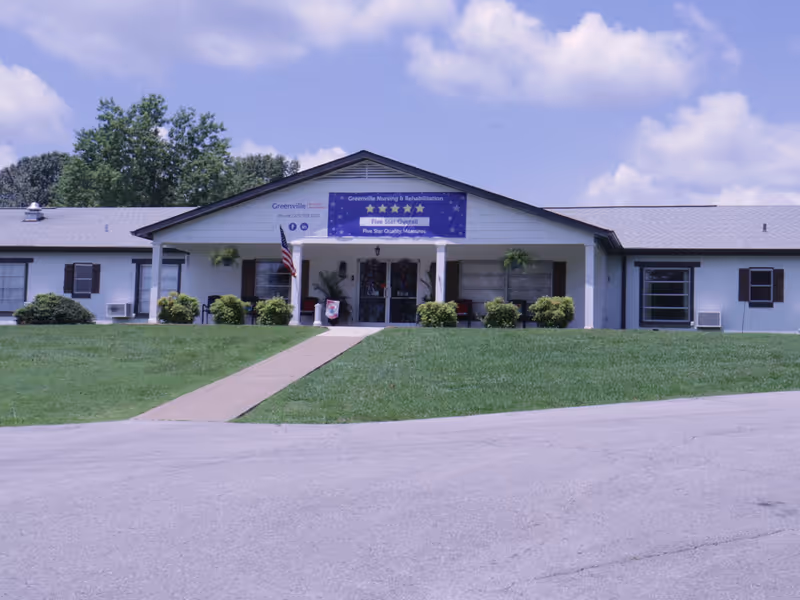 Front exterior view of Greenville Nursing and Rehabilitation building with a pathway leading to the entrance, green lawn, shrubs, an American flag, and a blue banner displaying a five-star rating above the entrance.