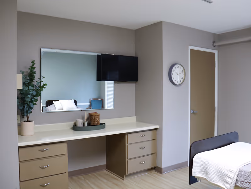 Furnished bedroom with a built-in desk and drawers, wall-mounted TV and mirror, clock on the wall, and a bed with a white blanket.
