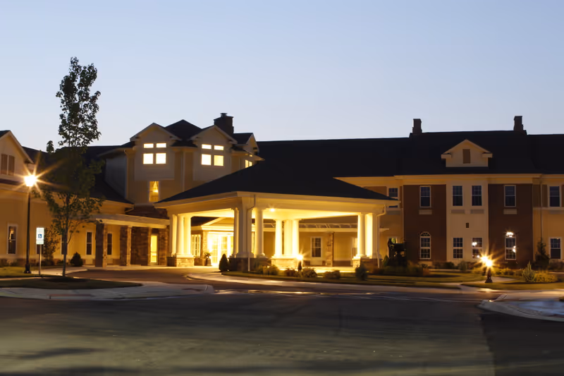 Exterior view of Fields of Florence: A Willow Ridge Senior Living Community building at dusk, with warm lights illuminating the entrance area featuring white columns and a covered driveway. The building has multiple windows and a mix of brick and siding exterior.