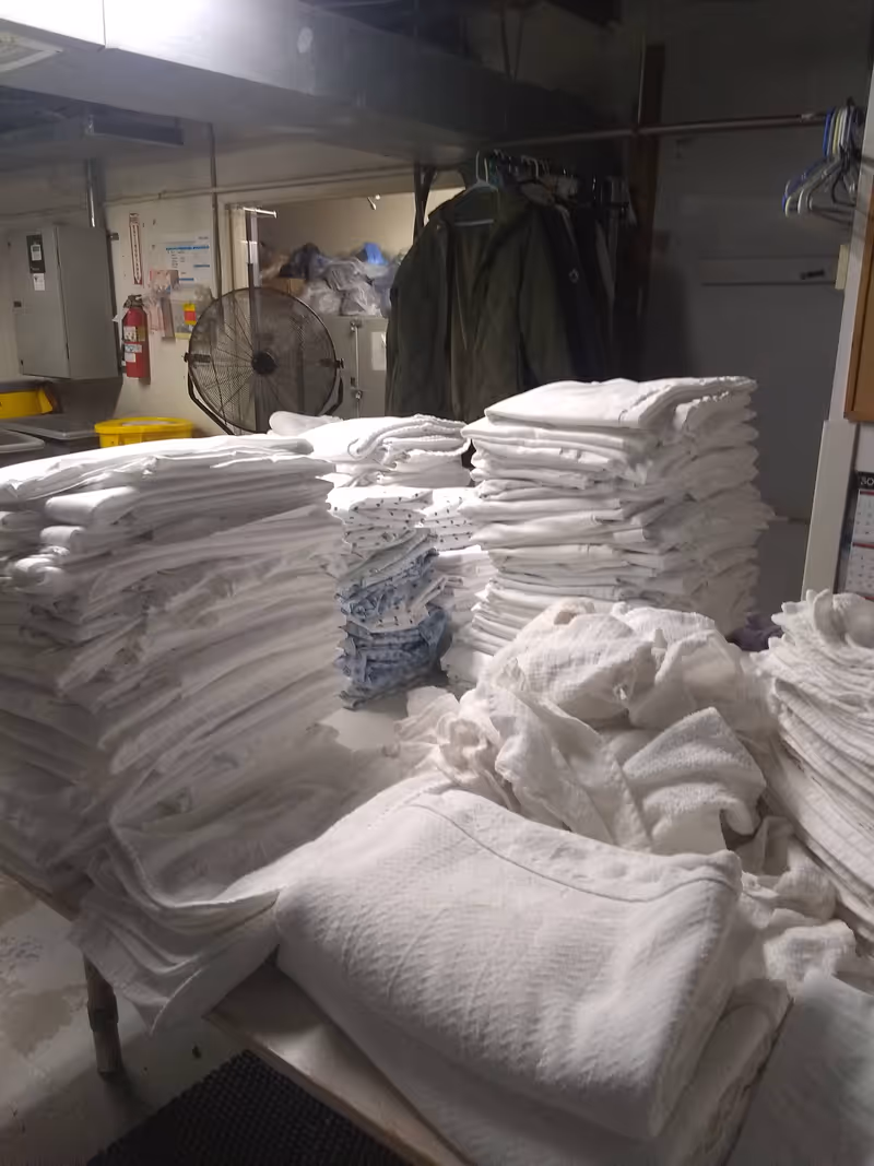 Stacks of folded white towels fill a laundry area in a facility's interior.