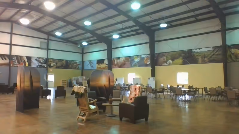 Large open interior space with high ceilings and exposed beams, featuring several seating areas with chairs and small tables. The walls have a mural strip with images of musical instruments, food, and nature scenes. The floor is polished concrete and the room is well-lit with ceiling lights.