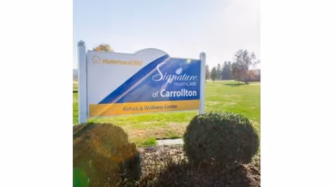 Outdoor lawn sign for "Signature HealthCARE of Carrollton Rehab & Wellness Center" with shrubs in front and a grassy field behind.