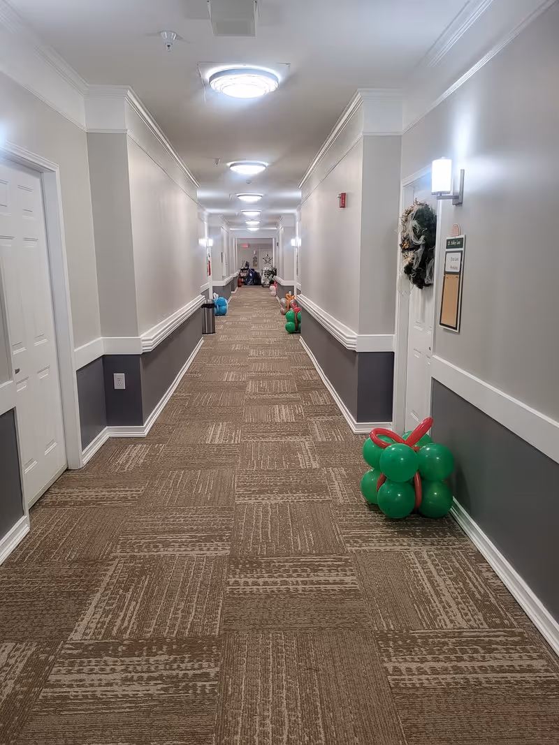 A long, well-lit hallway in a senior living facility with beige and gray walls, carpeted floor, and multiple closed doors on both sides. The hallway is decorated with clusters of colorful balloons and a wreath on one door.