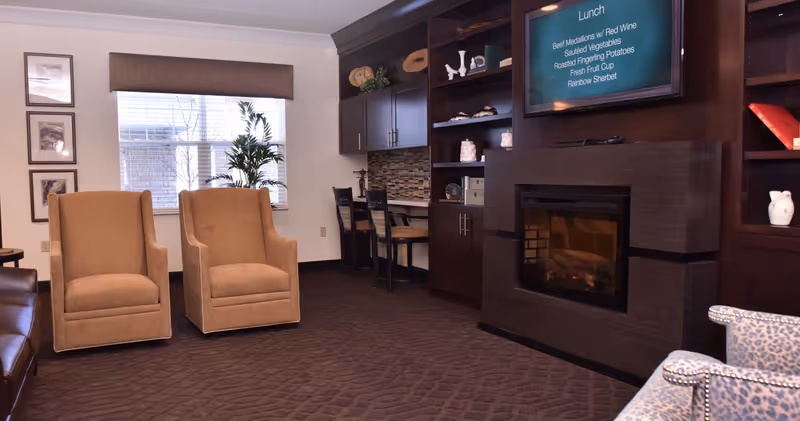 A cozy living room area with two beige armchairs, a window with blinds and a valance, a dark wooden built-in shelving unit with decorative items, a fireplace, and a wall-mounted TV displaying a lunch menu. There is also a small desk with two chairs and a patterned armchair partially visible.