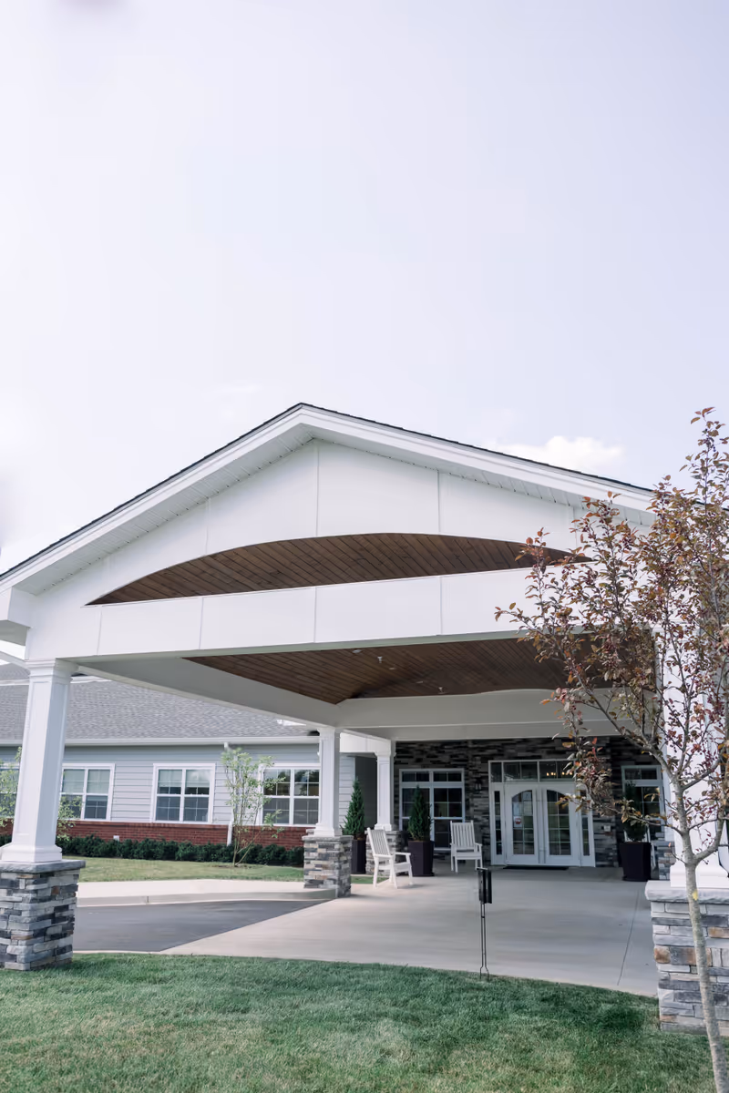 Covered porte-cochère and entrance of a single-story senior living building with white columns, stone pillars, and front seating.