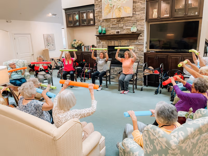 Residents and staff seated in a common living room circle lifting colorful foam noodles during a group exercise session.