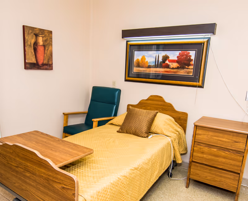 A small, simple bedroom with a single bed covered in a yellow quilt and a brown pillow. Next to the bed is a wooden nightstand with three drawers. A green armchair with wooden armrests is positioned beside the bed. On the wall above the bed is a framed landscape painting with autumn trees and a house, and another painting of a vase is hung on the adjacent wall.