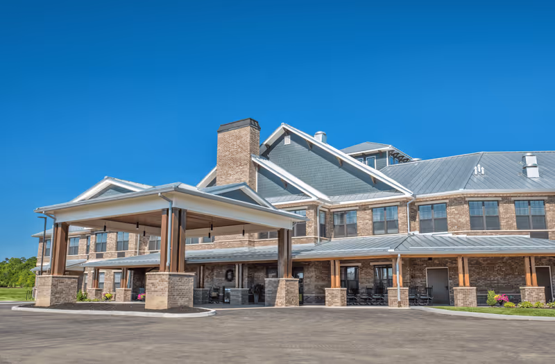 Exterior front view of a large senior living facility building with brick walls, multiple windows, and a covered entrance area under a clear blue sky.