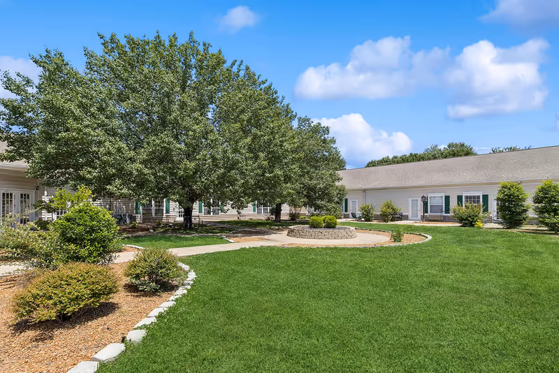A well-maintained outdoor courtyard area with green grass, a large leafy tree, shrubs, and a circular stone planter. The courtyard is surrounded by a single-story building with white siding and green shutters under a blue sky with some clouds.