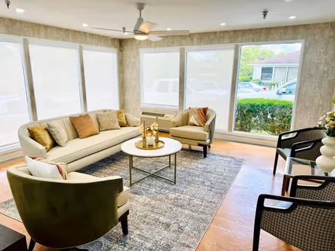 A bright and cozy living room with large windows covered by white blinds, a ceiling fan, a beige sectional sofa with multiple cushions, two armchairs, a round coffee table with decorative items, and a patterned area rug on a wooden floor.