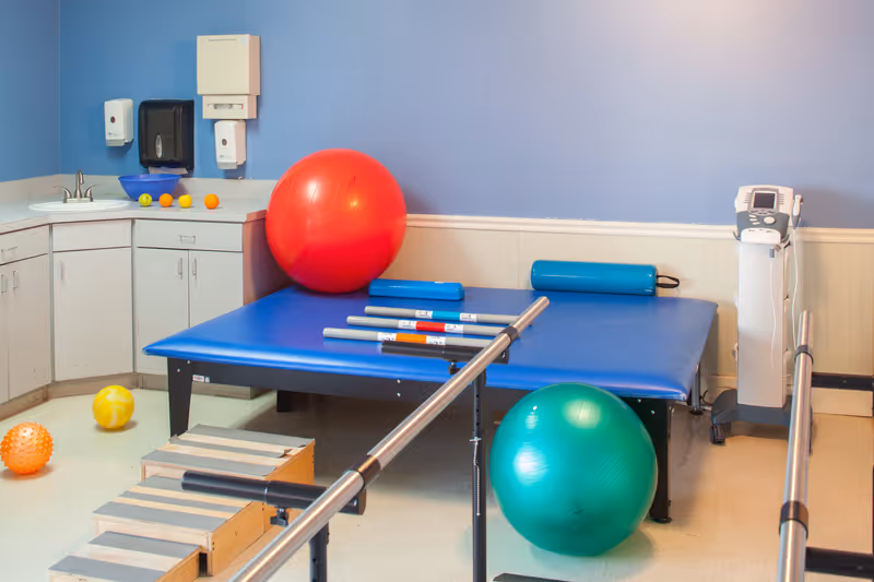 A physical therapy room with parallel bars, a treatment mat, large exercise balls, and cabinets against a blue wall.