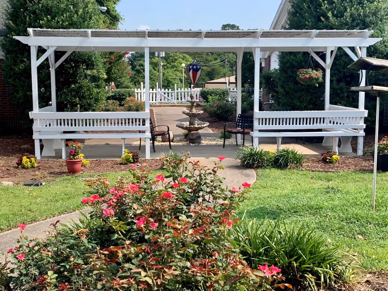 Outdoor garden area with a white wooden pergola structure featuring two white benches and two chairs underneath. There is a stone fountain with a patriotic hot air balloon decoration in the background, surrounded by green bushes and flowering plants. A curved concrete pathway leads through the garden with vibrant pink flowers in the foreground.