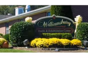 Outdoor view of a sign for Williamsburg Health & Rehabilitation Center surrounded by neatly trimmed bushes and yellow flowers, with part of a brick building visible in the background.