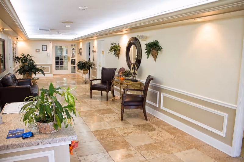 A well-lit interior hallway with beige tiled floors and cream-colored walls featuring decorative molding. The hallway includes two dark leather chairs and a small glass table with decorative items, a round wall mirror, and two wall-mounted plant holders with greenery. There are additional plants placed on a counter and in pots along the hallway. At the far end, there are glass double doors.