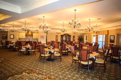 A spacious dining room with multiple round tables covered with patterned tablecloths and surrounded by wooden chairs with cushioned seats. The room features ornate chandeliers hanging from the ceiling, large windows with curtains, framed artwork on the walls, and a patterned carpeted floor.
