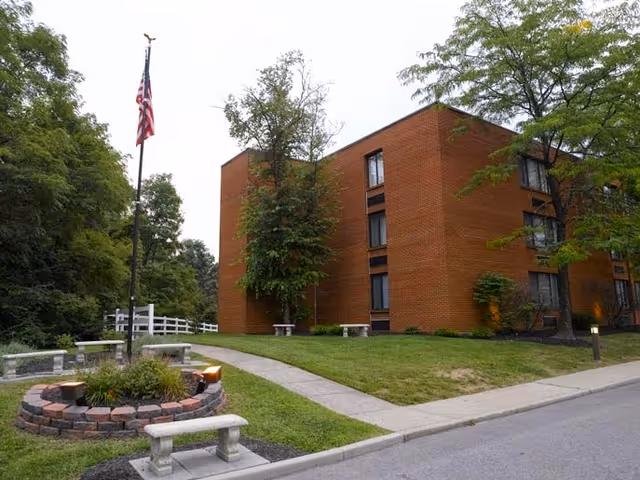 Front exterior of a brick multi-story senior living building with a flagpole, benches, walkway, and landscaped trees and lawn.