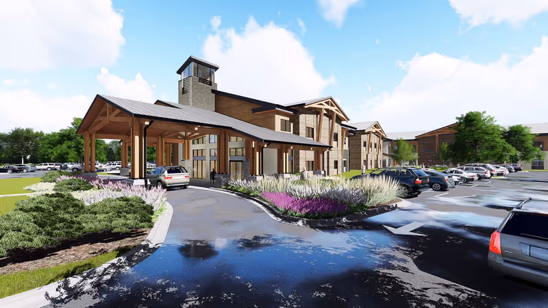 Exterior view of Boonespring Skilled Nursing Facility showing a large building with a covered entrance, surrounded by landscaped greenery and a parking lot with several parked cars under a partly cloudy sky.
