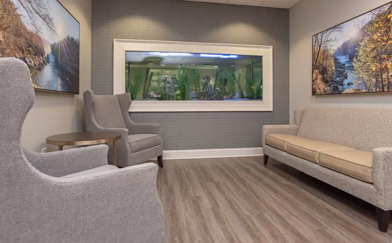 A cozy seating area with two gray armchairs and a gray sofa with beige cushions arranged around a small round wooden table. Behind the seating is a large built-in aquarium with green plants and decorative elements inside. The walls are decorated with two landscape paintings featuring nature scenes with trees and water. The floor is covered with wood-patterned vinyl or laminate.