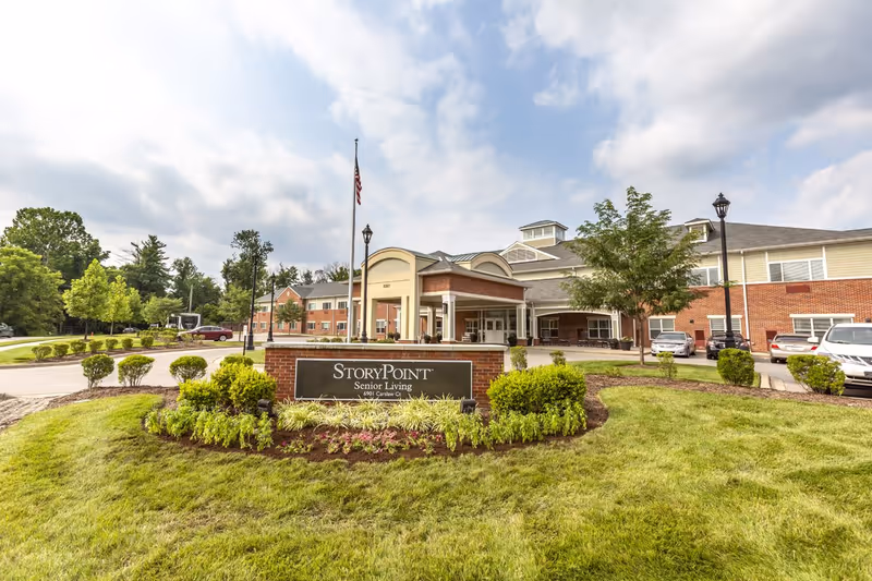 Exterior view of StoryPoint Senior Living facility with a brick and beige building, a covered entrance, an American flag on a flagpole, landscaped bushes and grass, and several parked cars under a partly cloudy sky.