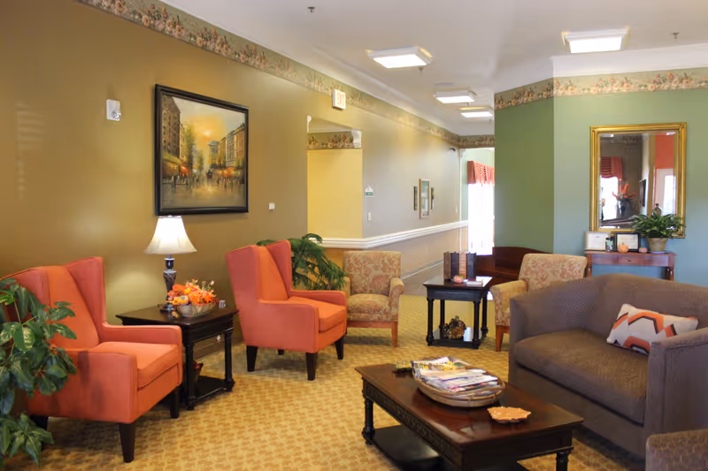 A cozy senior living facility common area with orange and patterned armchairs, a gray sofa with a decorative pillow, a wooden coffee table with magazines, a side table with a lamp and floral arrangement, a large framed painting on the wall, and a mirror above a small wooden console table with plants and decor.