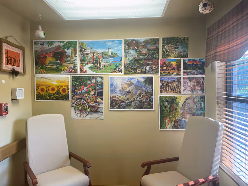 A cozy room with two white cushioned chairs with wooden armrests facing each other. The beige walls are decorated with multiple colorful paintings and pictures, including scenes of nature, animals, and landscapes. A window with blinds and a plaid valance allows natural light to enter the room. A small framed sign on the wall reads 'family'.