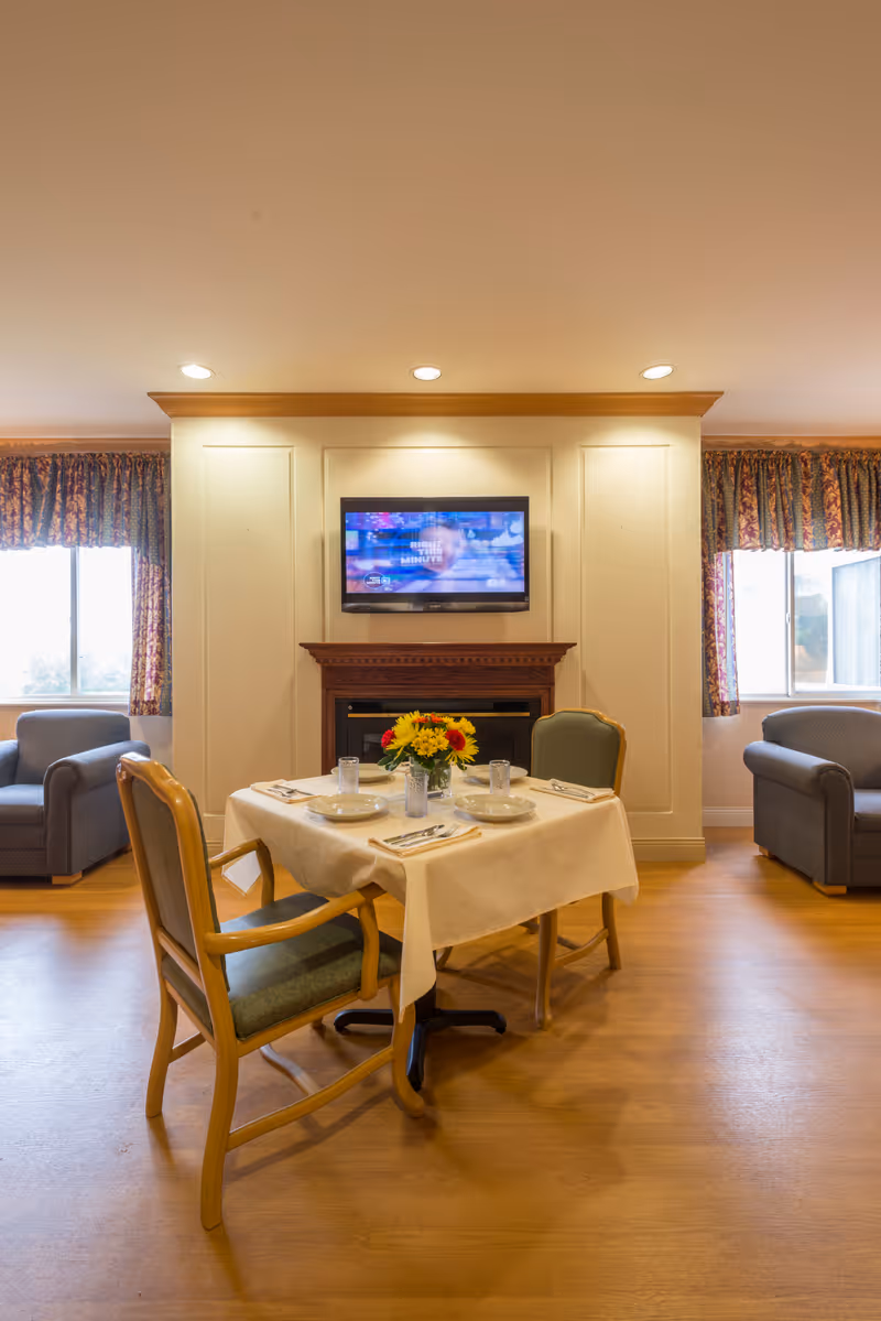 A cozy dining area with a small square table set for two, featuring a white tablecloth, plates, glasses, and silverware. A vase with sunflowers is placed in the center of the table. Behind the table is a wall with a mounted flat-screen TV above a wooden fireplace. Two armchairs are positioned near windows with patterned curtains on either side of the fireplace.