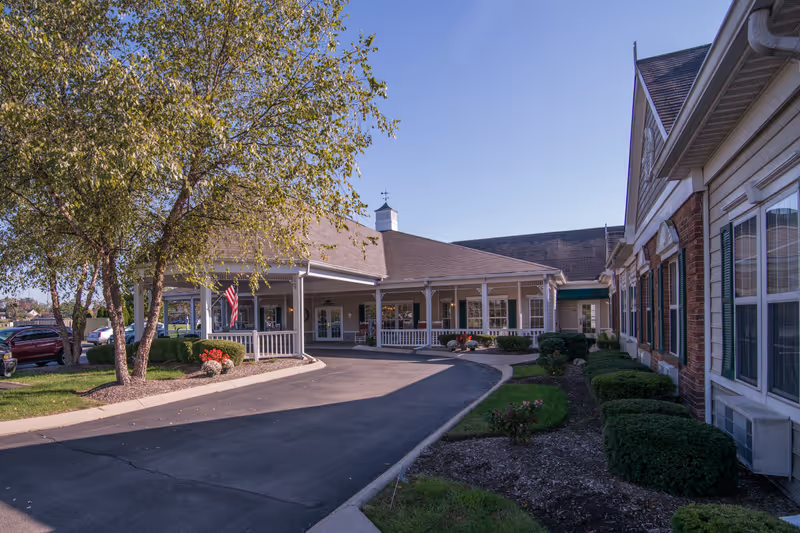 Exterior view of Morning Pointe of Owensboro facility showing a driveway leading to a covered entrance with white railings, surrounded by landscaped bushes and trees under a clear blue sky.