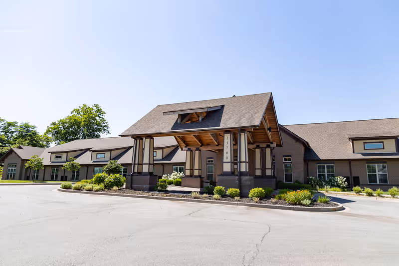 The front exterior of a single-story brick senior living building with a covered porte-cochere, landscaped beds, and a circular driveway.