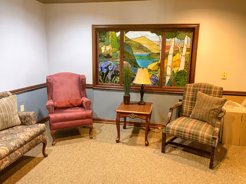 A cozy sitting area with three upholstered chairs arranged around a wooden side table holding a lamp and a potted plant. Behind the table is a large stained glass window depicting a colorful nature scene with trees, hills, and a river.