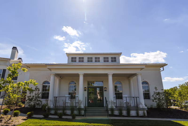 Front exterior view of a white building with large columns, green double doors, and several windows under a clear blue sky with some clouds. There are small bushes and trees planted around the building and a well-maintained lawn in front.