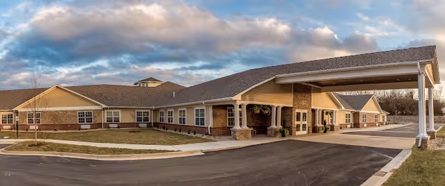 Exterior view of a single-story senior living facility building with a covered entrance driveway, beige siding, brick accents, and multiple windows under a partly cloudy sky.