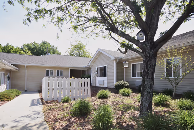 Courtyard of a single-story beige senior living building with a white picket fence, landscaped beds, and a tree.