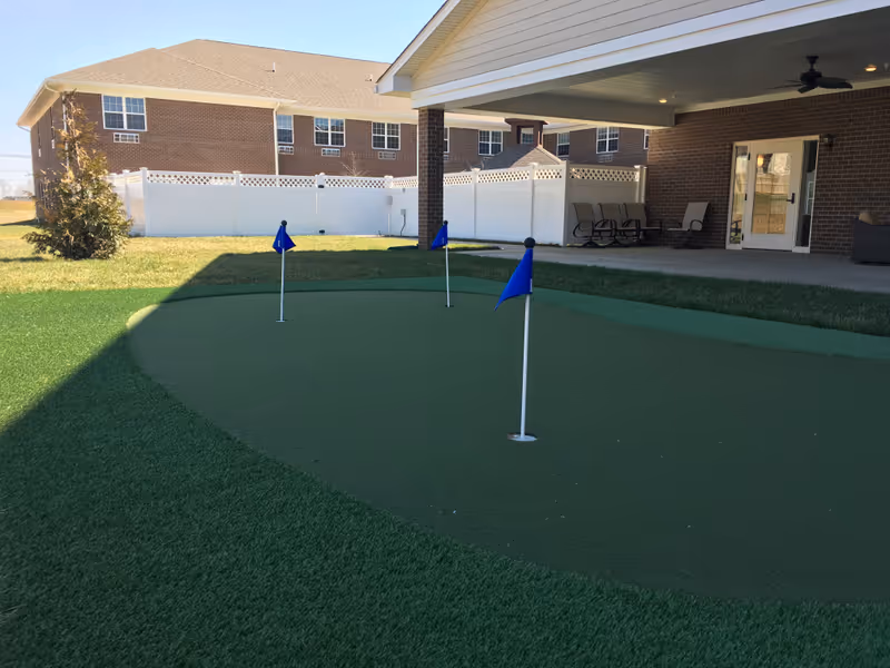 Outdoor putting green with three blue flags on a lawn area next to a covered patio with chairs and a ceiling fan. In the background, there is a brick building and a white fence.