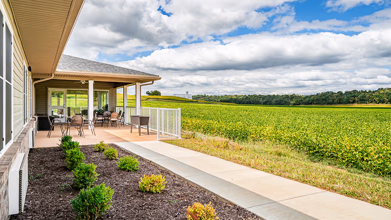 Covered patio area with several tables and chairs next to a building, overlooking a large green field under a partly cloudy sky.