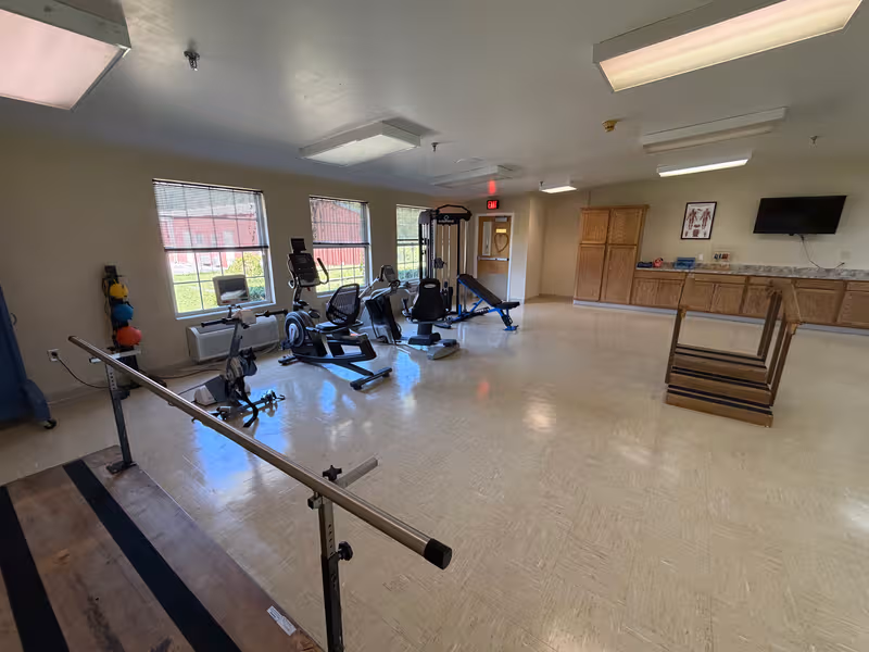 Senior living fitness room with cardio machines, weights, balance bars, and a wall-mounted TV.