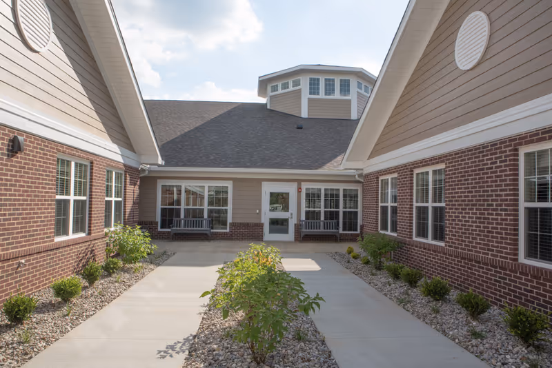 Central courtyard and entrance of a single-story brick building with walkways, benches, and landscaping under a partly cloudy sky.