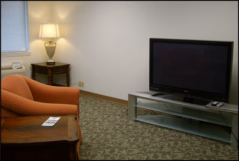 A small living room area with an orange patterned armchair, a wooden side table with a lamp, a flat-screen TV on a modern glass and metal TV stand, and a carpeted floor with a leafy pattern. There is a window with blinds and an air conditioning unit below it.