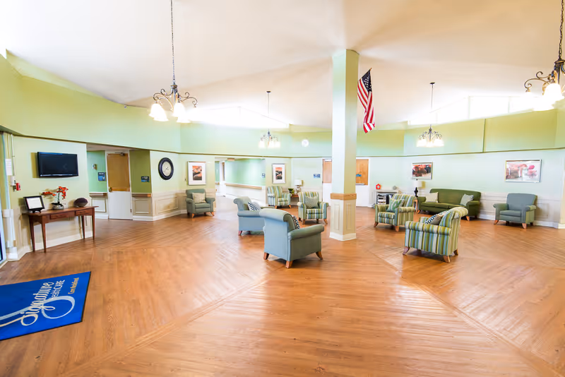 A spacious senior living facility common area with light green walls and wooden flooring. The room is furnished with several armchairs and sofas arranged in small groups. There are framed pictures on the walls, a wall-mounted TV, a clock, and a small table with decorative items. An American flag hangs from the ceiling, and multiple ceiling lights illuminate the space.