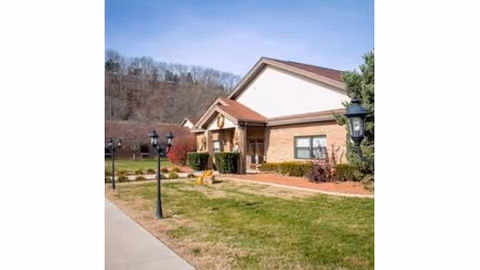 Exterior view of a single-story brick building with a gabled roof, surrounded by a grassy lawn and lamp posts along a sidewalk. Trees and hills are visible in the background under a clear blue sky.