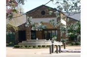 Exterior view of a senior living facility building with a brick facade, large windows, and a peaked roof. In front of the building is a circular stone fountain surrounded by a paved area and some landscaping with flowering trees and bushes.