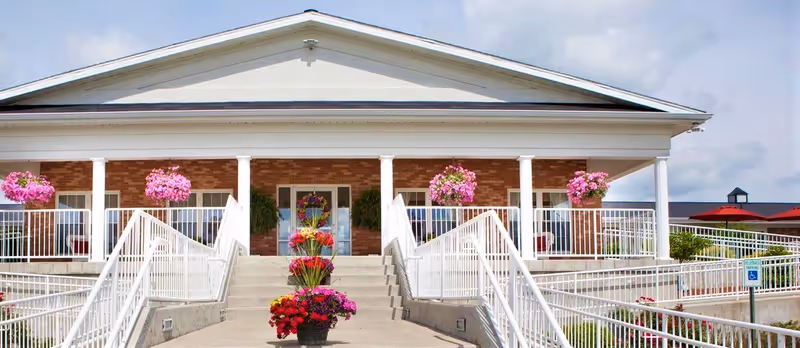 Front exterior view of a nursing and rehabilitation facility with a wide entrance ramp, white railings, brick walls, white columns, and hanging pink flower baskets. There are colorful flowers arranged in pots along the ramp.