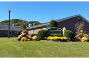 Brick single-story nursing home entrance with a manicured lawn, fall flower displays, and a 'Williamsburg' sign under a clear blue sky.
